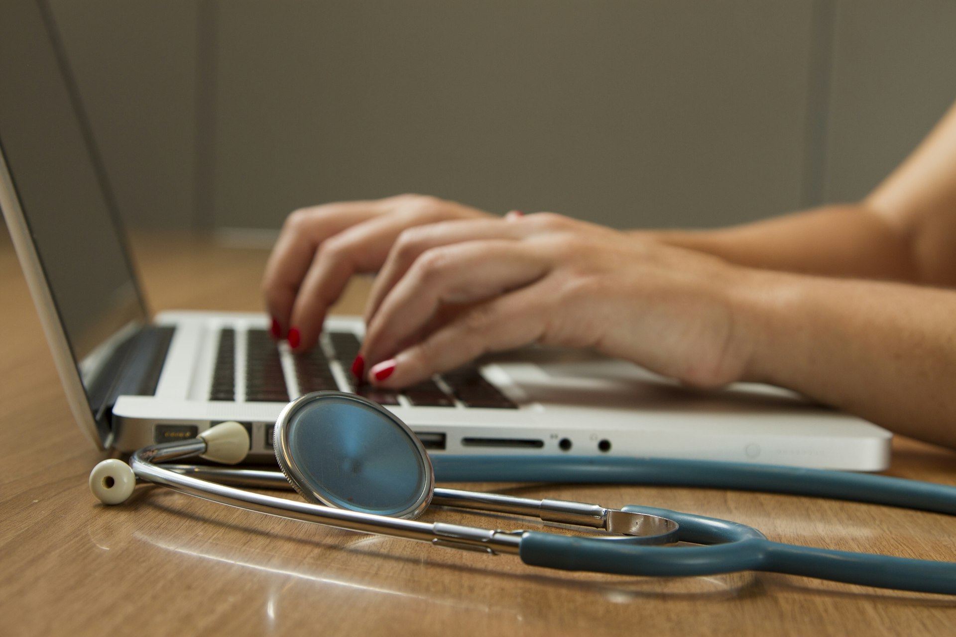Hands typing on a laptop beside a stethoscope on a desk
